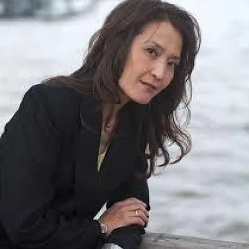 Eine Frau in einem schwarzen Blazer steht am Wasser. Ihr Blick ist nachdenklich und sie hat lange, lockige Haare.