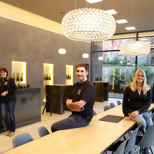 Ein modernes Weinlokal mit stilvollem Interieur und hellem Licht. Das Personal steht lächelnd im Raum, umgeben von Flaschen und einem großen Tisch.