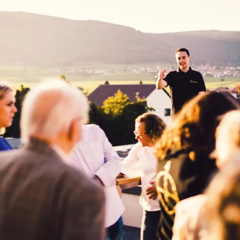 Eine Feier auf einer Dachterrasse mit Menschen, die sich versammeln und miteinander sprechen. Im Hintergrund ist eine malerische Landschaft und ein hügeliges Terrain zu sehen.