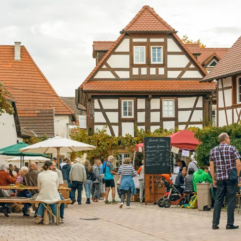 Eine lebhafte Straßenszene mit Menschen, die an Tischen sitzen und sich unterhalten. Im Hintergrund sind traditionelle Fachwerkhäuser zu sehen.