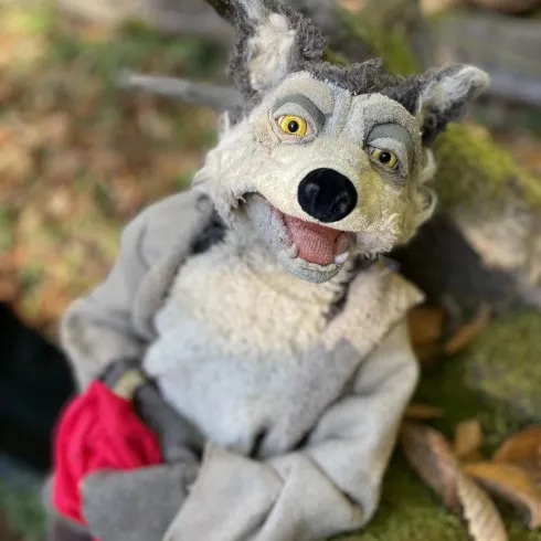 Eine Handpuppe eines Wolfes mit grauem Fell und einem freundlichen Gesichtsausdruck. Die Puppe hält ein rotes Tuch in der Hand und ist auf einem Baumstamm platziert.