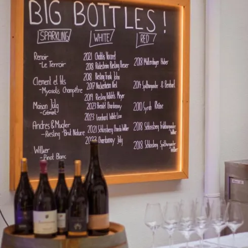 Eine Tafel mit der Aufschrift „BIG BOTTLES!“ zeigt eine Liste von Weinen. Darunter stehen verschiedene Weine auf einem Holzfass und Gläser.