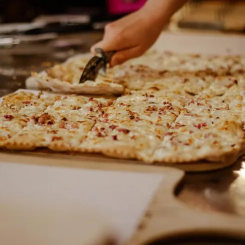 Ein frisch gebackenes Pizza, das in Stücke geschnitten wird. Im Hintergrund sind weitere Pizzastücke auf einer Holzplattform zu sehen.