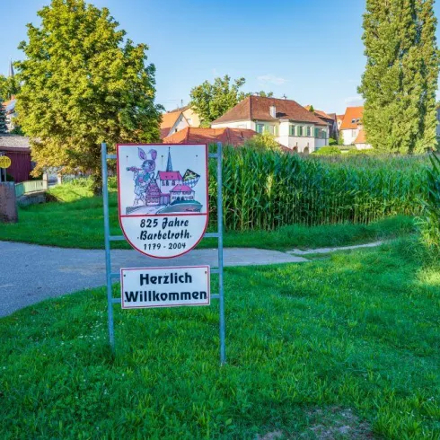 Ein Willkommensschild in einer ländlichen Umgebung. Im Hintergrund sind Häuser und hohe Pflanzen zu sehen.