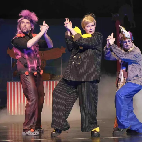 Eine Gruppe von fünf Schauspielern in bunten Kostümen steht auf einer Bühne. Sie posieren mit lustigen Gesten und wirken fröhlich und energiegeladen.