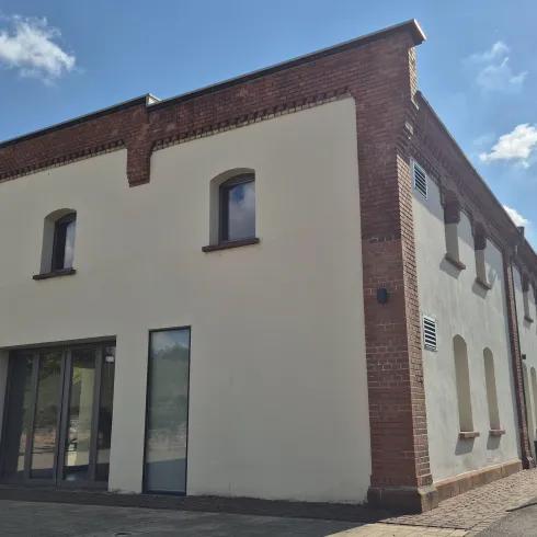 Ein modernes Gebäude mit einer weißen Fassade und einigen Fenstern. Im Hintergrund sind Bäume und ein blauer Himmel mit Wolken zu sehen.