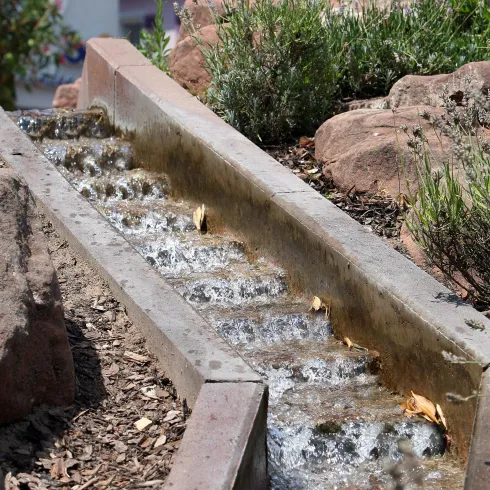 Ein Wasserlauf aus Beton mit fließendem Wasser in einem begrünten Bereich. Im Hintergrund sind orangefarbene Dächer und eine Statue sichtbar.