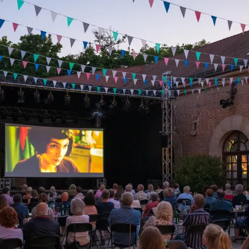 Ein Freiluftkino mit vielen Zuschauern, die auf eine große Leinwand schauen. Bunte Fahnen sind über den Platz gespannt, während die Dämmerung einsetzt.