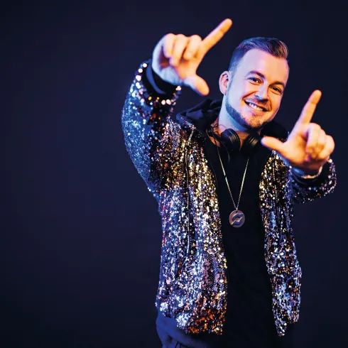 Ein junger Mann trägt eine glitzernde Jacke und Kopfhörer. Er lächelt und macht mit seinen Händen ein Zeichen.
