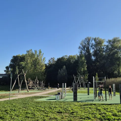 Ein Spielplatz mit verschiedenen Klettergeräten und Schaukeln in einem grünen Park. Der Himmel ist klar und die Sonne scheint.