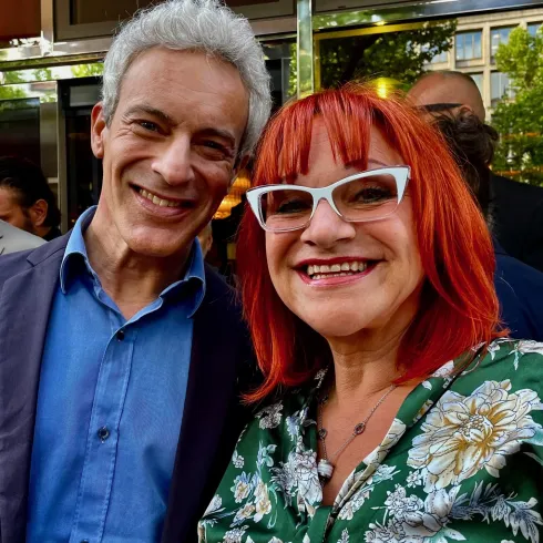 Ein lächelndes Paar steht vor einem Gebäude. Die Frau hat rote Haare und eine auffällige Brille, während der Mann einen blauen Hemd trägt.