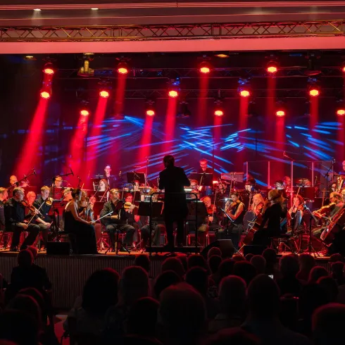 Ein Orchester spielt auf einer Bühne mit bunten Lichtstrahlen. Zwei Sänger stehen im Vordergrund und präsentieren eine beeindruckende Darbietung.