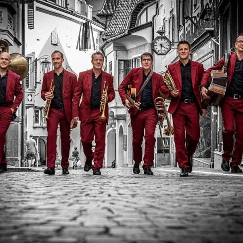 Eine Gruppe von sechs Musikern in roten Anzügen geht fröhlich eine historische Straße entlang. Sie halten verschiedene Musikinstrumente in den Händen und haben einen energiegeladenen Ausdruck.
