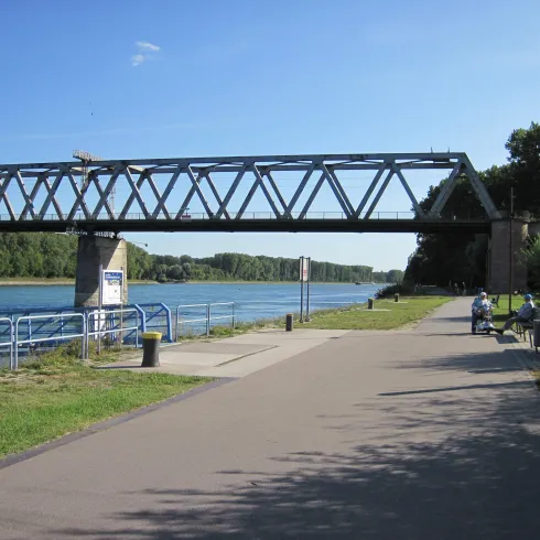 Eine Fußgängerbrücke über einen Fluss, umgeben von grünem Gras und Bäumen. Auf dem Weg sitzen Menschen und genießen die Aussicht.