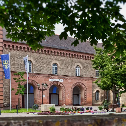 Ein historisches Gebäude mit roten Backsteinmauern und schönen Fenstern. Darum herum sind grüne Bäume und bunte Blumen.