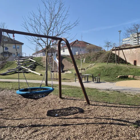 Ein Spielplatz mit einer Schaukel und einem großen Sandbereich. Im Hintergrund sind einige Bäume und Wohngebäude zu sehen.