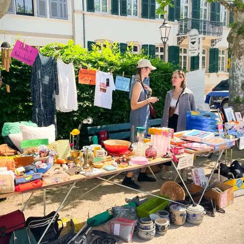 Ein Flohmarkt mit verschiedenen Ständen und Angeboten. Zwei Frauen unterhalten sich vor einer Auswahl an Second-Hand-Artikeln.