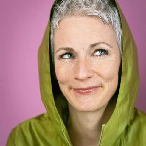 Eine Frau mit grauen Haaren trägt eine grüne Jacke mit Kapuze. Sie hat ein freundliches Lächeln und einen verspielten Blick vor einem rosa Hintergrund.