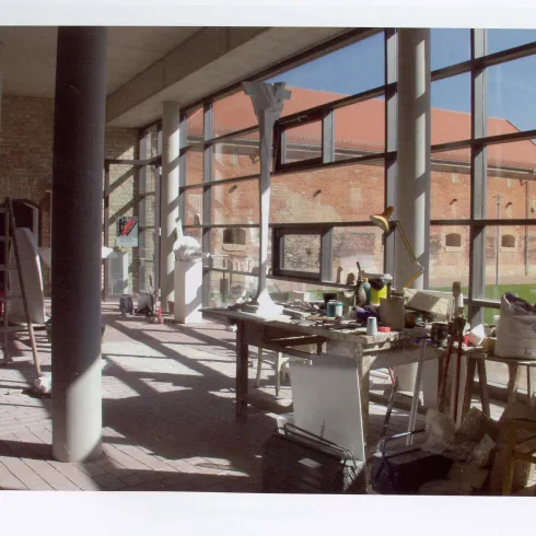 Ein helles Atelier mit großen Fenstern und einem Blick auf eine Backsteinmauer. Auf einem Tisch sind verschiedene Werkzeuge und Materialien verteilt.
