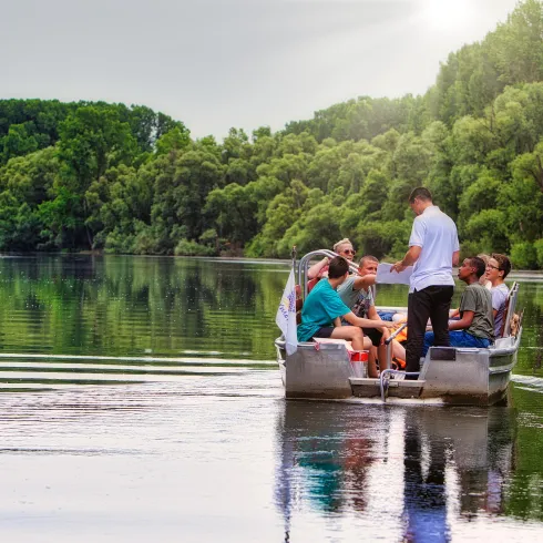 Eine Gruppe von Personen sitzt in einem Boot auf einem ruhigen Fluss. Umgeben von grünen Bäumen und einer friedlichen Landschaft genießen sie die Natur.