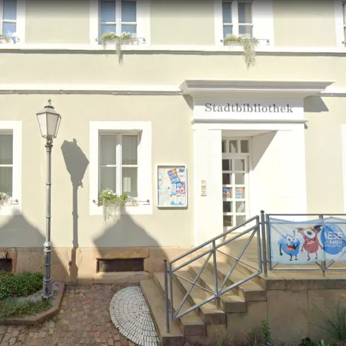Eine Stadtbibliothek mit einem einladenden Eingangsbereich. Vor der Bibliothek steht eine Laterne und ein buntes Banner.