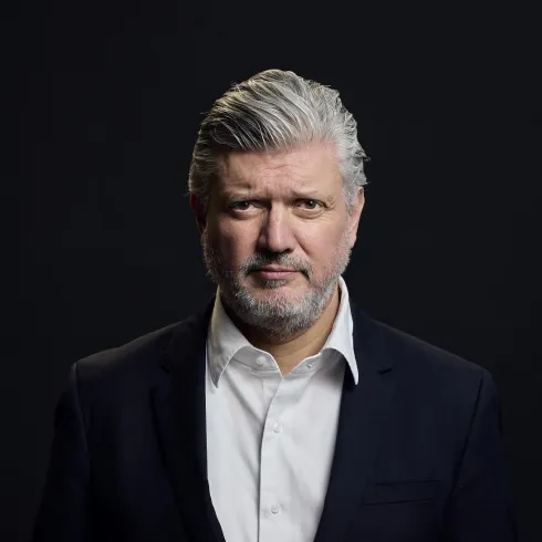 Ein reifer Mann mit grauen Haaren und einem Bart steht vor einem dunklen Hintergrund. Er trägt ein schwarzes Jackett und ein weißes Hemd.