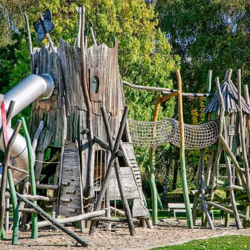 Ein kreativer Spielplatz aus Holz mit einer Rutsche und einem Klettergerüst. Die Struktur sieht wie ein Baumstamm aus und bietet viele Möglichkeiten zum Spielen.