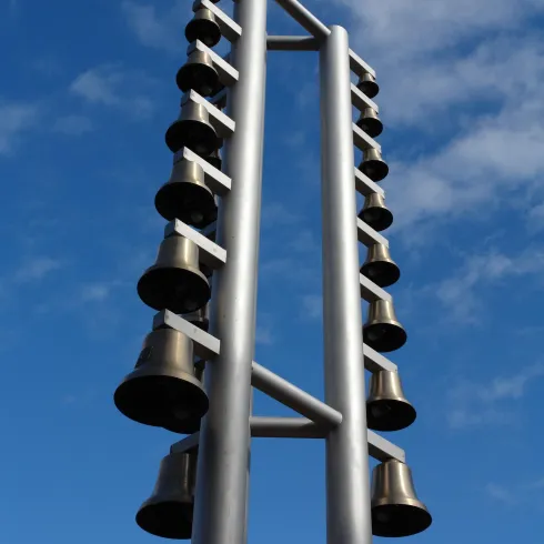 Ein echter Glockenbaum aus Metall mit mehreren hängenden Glocken. Der Himmel ist blau mit einigen Wolken.