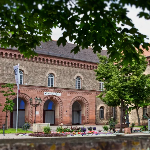 Ein historisches Gebäude mit einer schönen Fassade und bunten Blumen im Vordergrund. Bäume umgeben den Platz und verleihen eine grüne Atmosphäre.