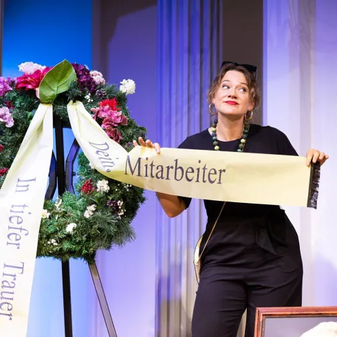 Eine Schauspielerin zeigt ein Trauergesteck mit einem Banner. Sie steht im Mittelpunkt der Bühne und wirkt nachdenklich.