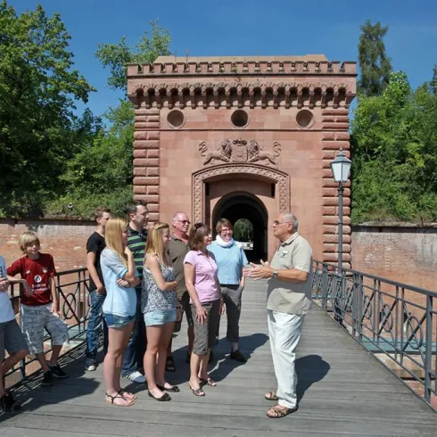 Eine Gruppe von Menschen steht auf einer Holzbrücke vor einem historischen Gebäude. Es ist ein sonniger Tag und die Umgebung ist grün und einladend.