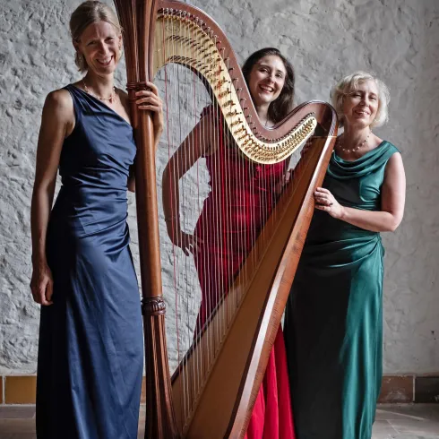 Drei Frauen stehen lächelnd vor einer Harfe. Sie tragen elegante Kleider in verschiedenen Farben.