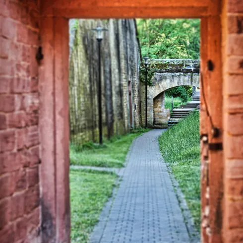 Ein schmaler Weg, der durch einen Torbogen führt. Auf beiden Seiten sind grüne Wiesen und historische Mauern zu sehen.