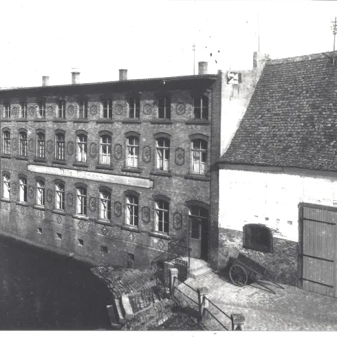 Ein historisches Gebäude mit mehreren Fenstern vorn, umgeben von Wasser und alten Zäunen. Im Hintergrund ist ein angrenzendes Gebäude mit einem Schuppen zu sehen.