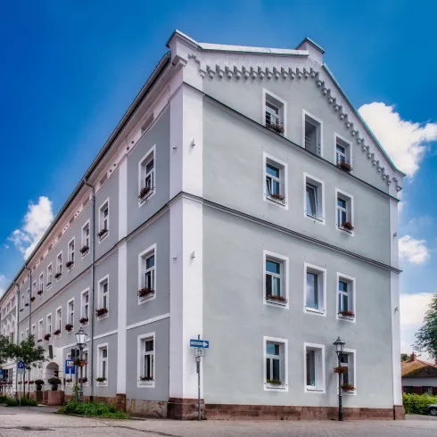 Ein graues Wohngebäude mit mehreren Fenstern und kleinen Balkonen. Im Hintergrund sind Bäume und ein blauer Himmel zu sehen.