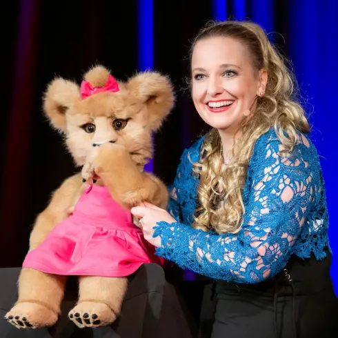 Eine lächelnde Frau präsentiert eine Stofftierpuppe in einem pinken Kleid. Im Hintergrund sind bunte Lichter zu sehen.