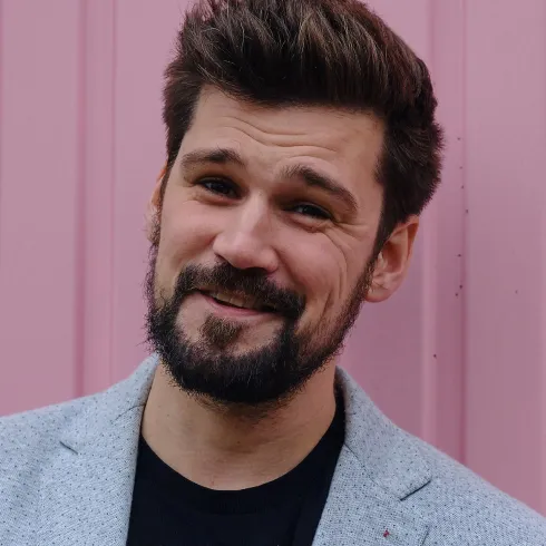 Ein junger Mann mit einem lächelnden Gesicht steht vor einer pinken Wand. Er trägt einen grauen Blazer und ein schwarzes T-Shirt.