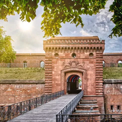Ein historisches Tor mit einer Holzbrücke führt in eine Festungsanlage. Die Umgebung ist grün und sonnig, was eine einladende Atmosphäre schafft.