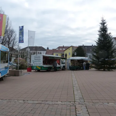 Ein Platz mit mehreren Food Trucks und einem Weihnachtsbaum. Im Hintergrund sind bunte Gebäude und Fahnen zu sehen.