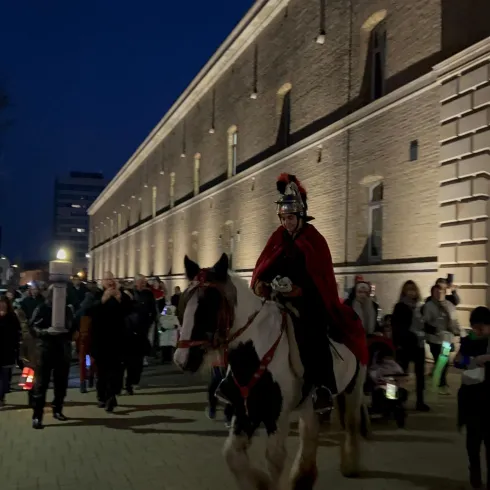 Eine Gruppe von Menschen mit einem Ritt auf einem Pferd. Der Reiter trägt einen roten Umhang und die Szene spielt sich in der Dämmerung vor einer historischen Mauer ab.
