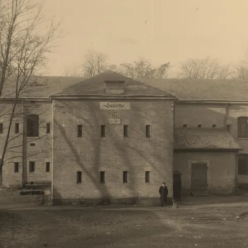 Ein historisches Gebäude umgeben von Bäumen. Zwei Personen sind im Vordergrund zu sehen.