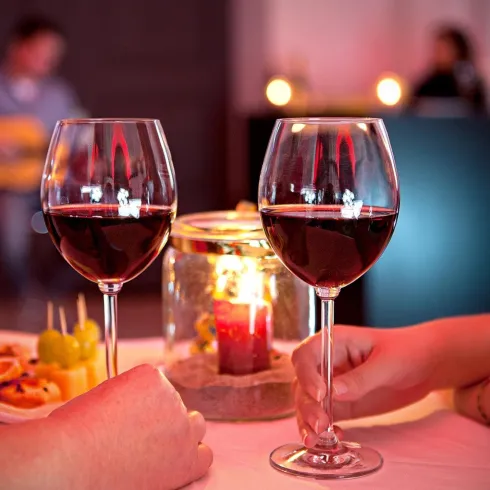 Zwei Gläser Rotwein stehen auf einem Tisch, umgeben von einer Auswahl an Snacks. Im Hintergrund spielt ein Musiker Gitarre und es herrscht eine romantische Atmosphäre.
