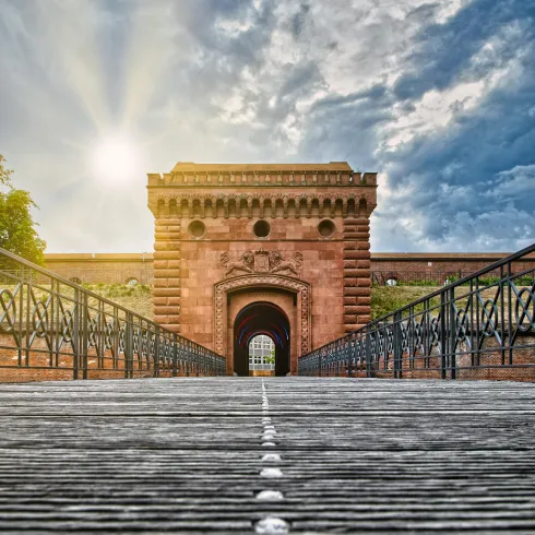 Ein beeindruckendes historisches Tor mit einer breiten Holzbrücke davor. Der Himmel ist blau mit einigen Wolken und der Sonnenstrahl beleuchtet die Szenerie.