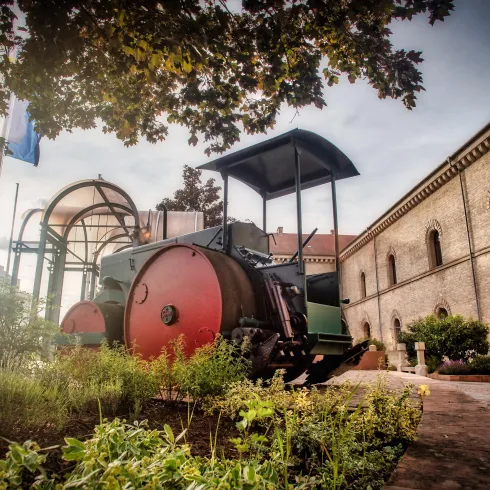 Ein historischer Straßenwalzen mit roten Walzen steht vor einem alten Gebäude. Im Hintergrund sind einige Bäume und Flaggen zu sehen.