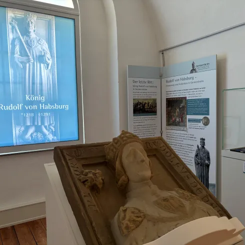 Eine Skulptur von König Rudolf von Habsburg steht in einem Museum. Im Hintergrund sind Informationsplakate und ein Fenster mit einer Darstellung des Königs zu sehen.