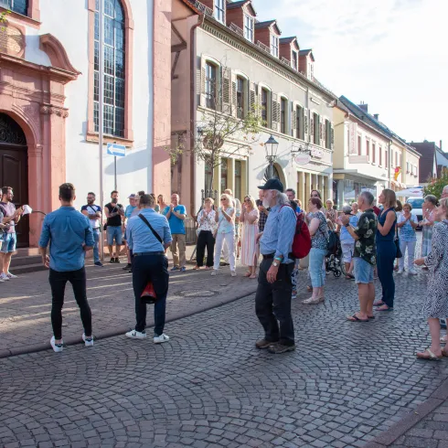 Eine Gruppe von Menschen versammelt sich in einer malerischen Straße, während ein Musiker auf der Gitarre spielt. Die Atmosphäre ist lebhaft und freundlich.