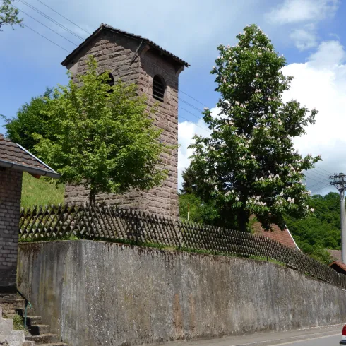 Ein kleiner, steinerner Uhrturm steht in einer ländlichen Umgebung. Neben dem Turm wachsen grüne Bäume und die Straße verläuft in der Nähe.