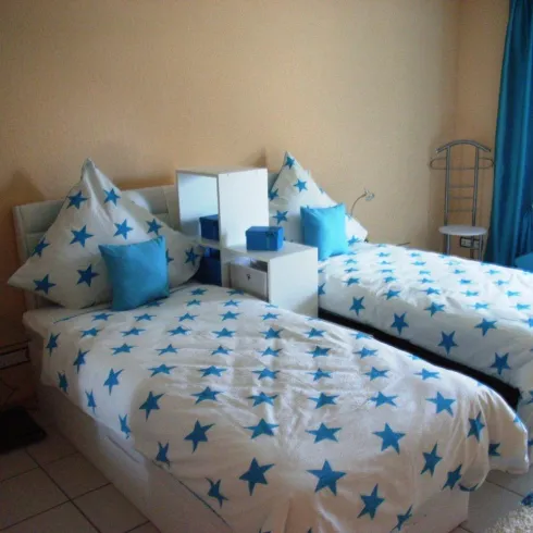 Ein gemütliches Schlafzimmer mit zwei Einzelbetten, die mit Sternenmuster und blauen Kissen dekoriert sind. Der Raum hat helle Wände und einen großen Fenstervorhang in Blau.