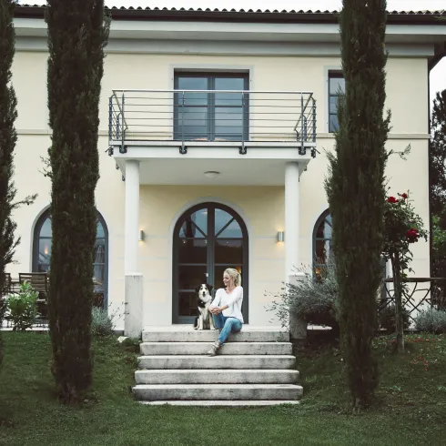Ein schönes Haus mit zwei Säulen und großen Fenstern. Eine Frau sitzt auf den Treppen und ein kleiner Hund ist bei ihr.