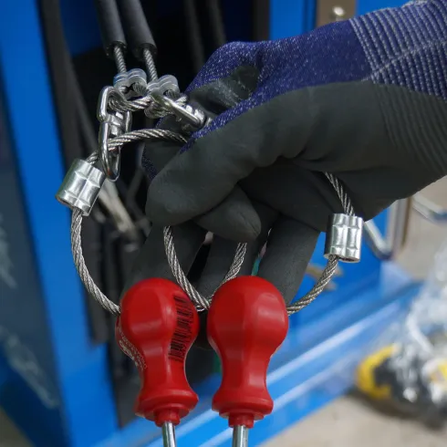 Eine Hand hält zwei rote Schraubenzieher an Drahtseilen. Im Hintergrund sind weitere Werkzeuge zu sehen.